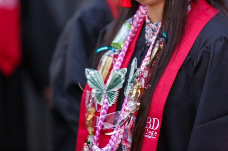 A young woman celebrates her graduation from CBD College, wearing a traditional graduation gown. Adorning her neck is a necklace embellished with delicate butterfly charms, adding a touch of elegance and personal style to her academic achievement.