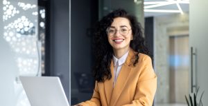 A woman specializing in health sciences sits at a desk, a computer before her, radiating a warm smile. Her demeanor reflects a positive and engaged presence as she likely navigates the intersection of technology and health sciences in her work or studies.