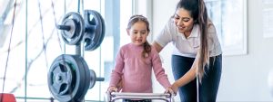 An occupational therapy technician assists a seven-year-old girl with a fracture in her lower extremities as she navigates rehabilitation equipment within the therapy room. The technician offers supportive guidance and encouragement, helping the girl regain mobility and independence during her recovery journey.