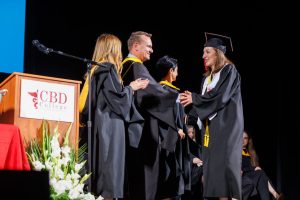 A female CBD College graduate receiving her certificate from a professor on stage during the graduation ceremony.