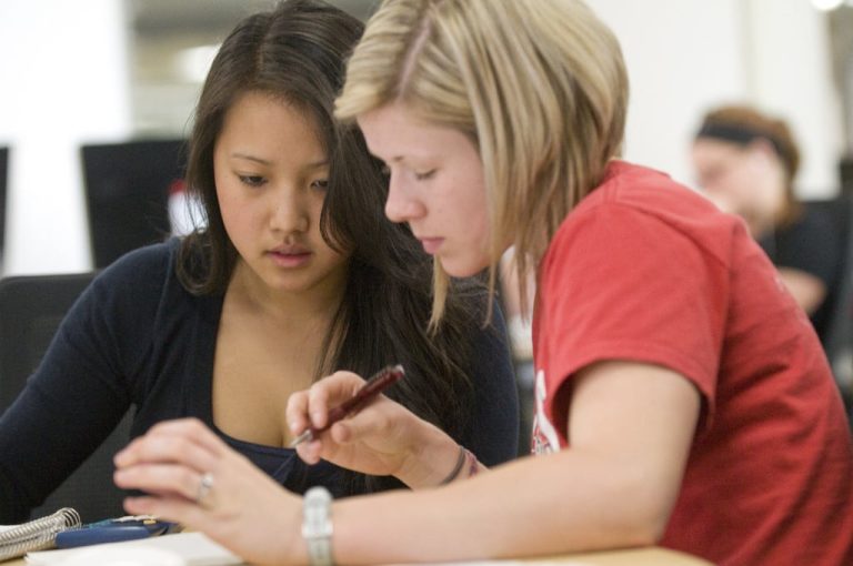 Two female students studying together at a desk, with textbooks and notebooks open, engaged in collaborative learning.