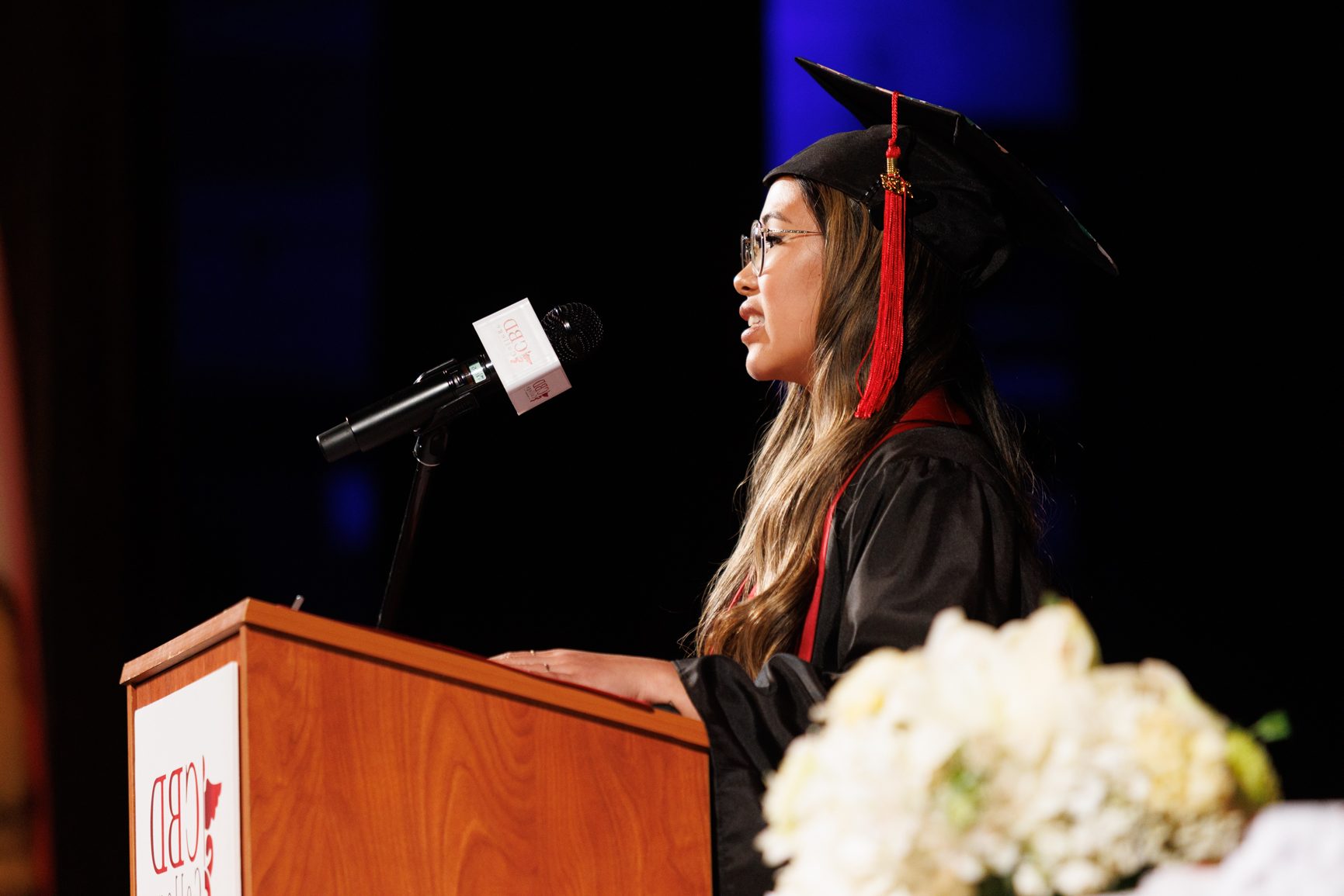 A CBD College graduate, a young woman, stands confidently on the graduation stage, adorned in a graduation robe and cap. In front of her, a table holds a microphone, symbolizing her commencement speech, as she addresses the audience with pride and accomplishment.