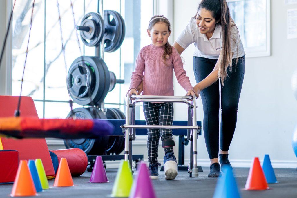 An occupational therapy technician assists a seven-year-old girl with a fracture in her lower extremities as she navigates rehabilitation equipment within the therapy room. The technician offers supportive guidance and encouragement, helping the girl regain mobility and independence during her recovery journey.