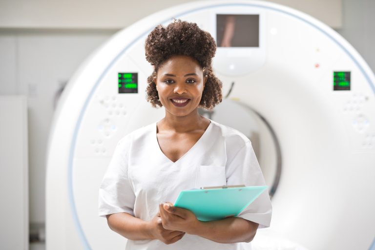 A female doctor standing in a medical examination room against an MRI scan, holding a clipboard, and smiling at the camera.