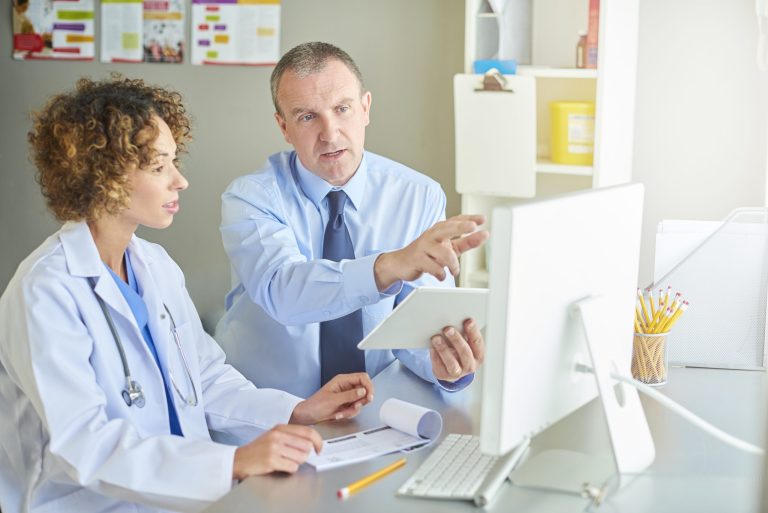 An administrator sits with a female doctor, leading a presentation while referring to the computer screen on the desk.