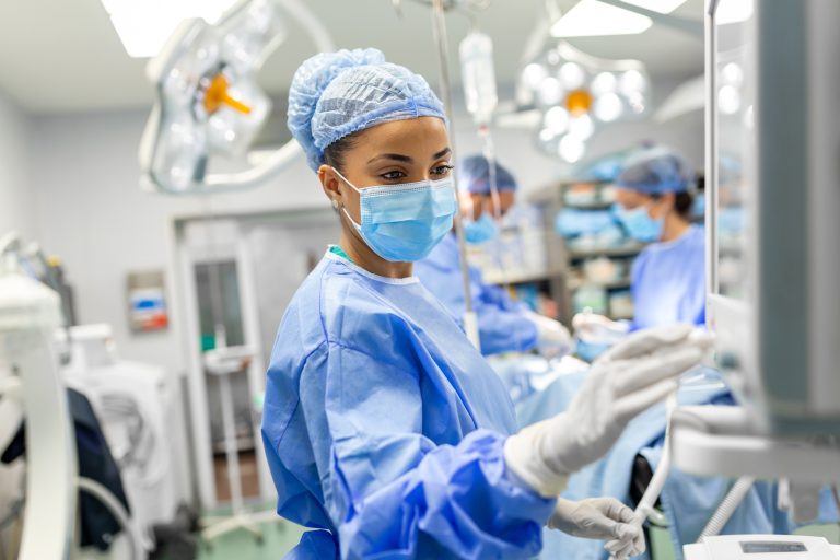 A young female doctor wearing scrubs and a protective face mask prepares an anesthesia machine before an operation.
