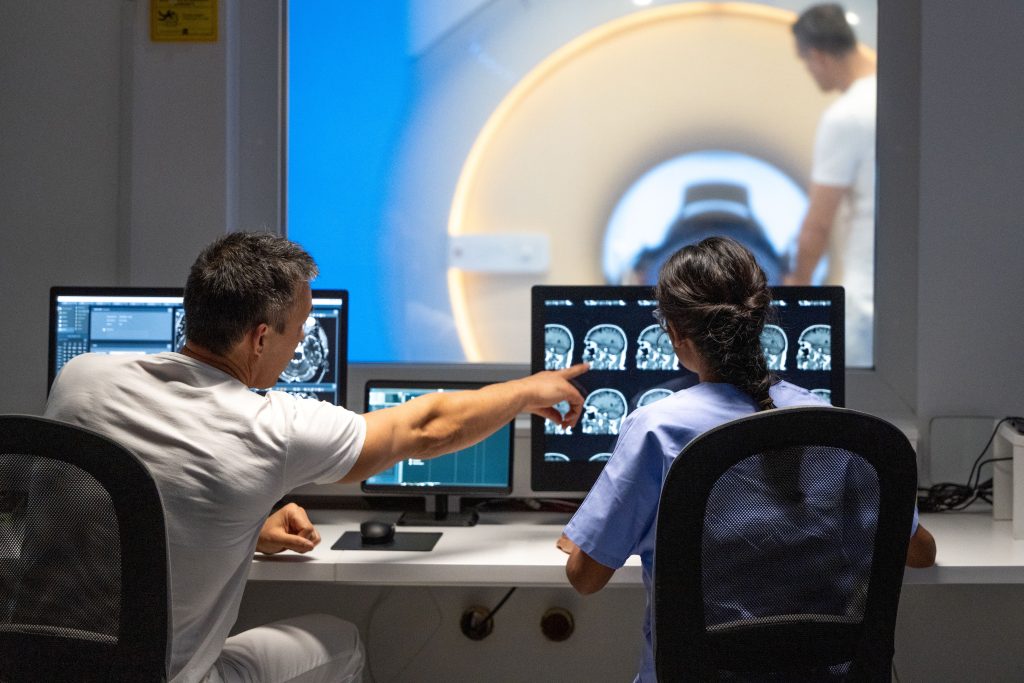 Two doctors are inside an MRI buffer room, discussing a special brain X-ray image displayed in front of them. In the MRI room, an imaging technician operates the machine while a patient undergoes imaging. The doctors examine the results, collaborating to provide optimal patient care.